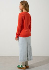 Roter gestrickter Pullover, hellblauer Jeansrock mit Schlitz, grüne Clogs und hellfarbige Socken. Einfaches Design, lässige Passform und strukturierter Stoff.