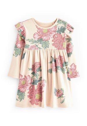 Jeune enfant portant une robe rose à fleurs avec des épaules volantées, des chaussettes blanches à volants et des baskets roses à scratch, debout devant un fond vert.