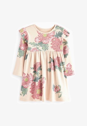 Jeune enfant portant une robe rose à fleurs avec des épaules volantées, des chaussettes blanches à volants et des baskets roses à scratch, debout devant un fond vert.