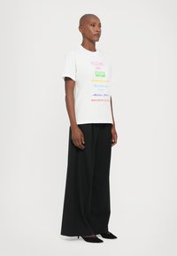 White t-shirt featuring multicolored text logos, paired with loose black trousers and black pointed flats, against a plain background.
