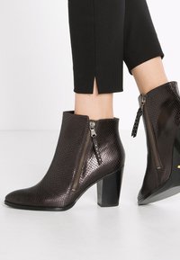 Stivali alla caviglia in pelle di serpente marrone scuro, con una finitura testurizzata, punta appuntita e tacco a blocco di altezza media; presenta cerniere su entrambi i lati.