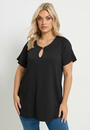 Donna bionda che indossa una maglia nera a maniche corte con una piccola apertura frontale a forma di chiave e una fibbia a forma di cuore, abbinata a jeans blu, su uno sfondo semplice.