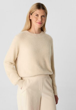 Frau mit langen blonden Haaren, die einen beigen, flauschigen Pullover und hellbeige, hochtaillierte Hosen trägt, steht mit einer Hand in der Tasche.