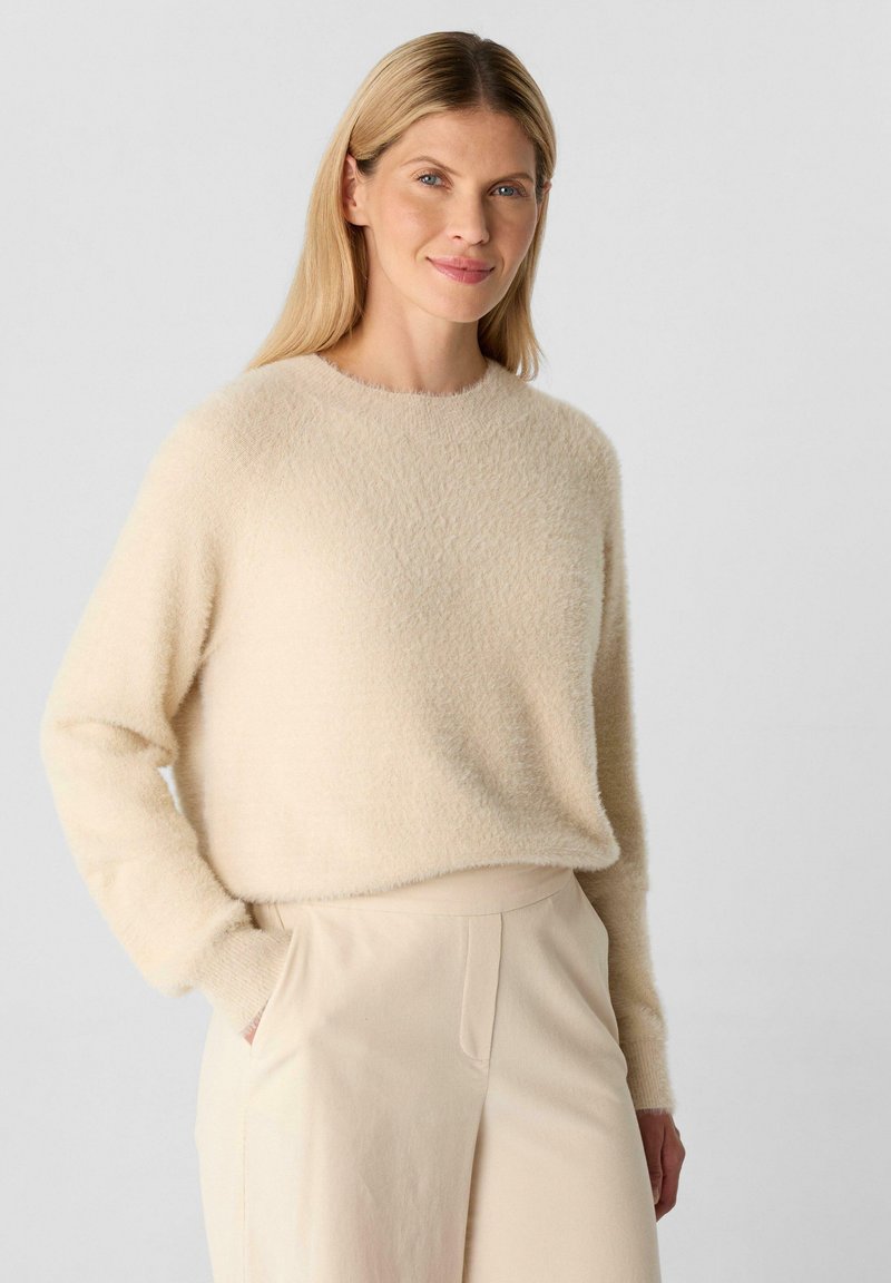 Frau mit langen blonden Haaren, die einen beigen, flauschigen Pullover und hellbeige, hochtaillierte Hosen trägt, steht mit einer Hand in der Tasche.