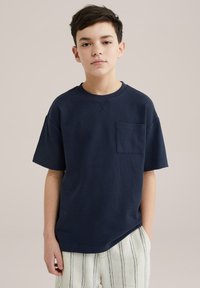 Temno modra majica s kratkimi rokavi in krožnim ovratnikom, z sprednjo žepno in teksturiranim vzorcem, v kombinaciji s kremnato prugastimi hlačami.