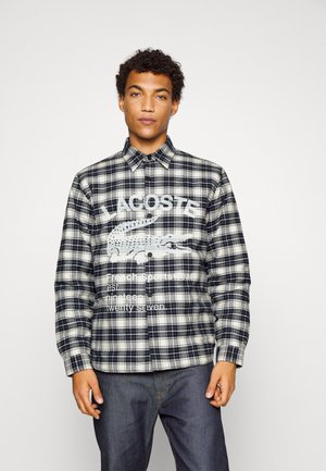 Chemise à carreaux à boutons en bleu marine, blanc et gris avec un grand logo imprimé de crocodile et du texte sur la poitrine. Fabriquée en tissu tissé.