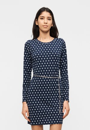 Γυναίκα που φοράει μακρυμάνικο φόρεμα σκούρου μπλε με λευκές βούλες και ασημένια ζώνη αλυσίδα, στέκεται μπροστά σε απλό ανοιχτό φόντο.