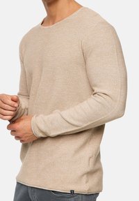 Beige pitkähihaine neulepusero, jossa on pyöreä pääntie, teksturoitu kangas ja hienovaraiset ompeluyksityiskohdat. Ominaisuuksiin kuuluu istuvat hihansuut ja helma.