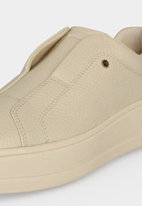 Μπεζ υφασμάτινο δερμάτινο slip-on sneaker με παχιά σόλα και διακοσμητικές ραφές, που δείχνει την επάνω πλευρά και τη λεπτομέρεια μεταλλικών οπών.