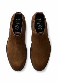 Brązowe zamszowe buty wsuwane z elastycznymi panelami po bokach i teksturowanymi ciemnymi gumowymi podeszwami. Logo BOSS widoczne na wkładkach. Wyprodukowano we Włoszech.