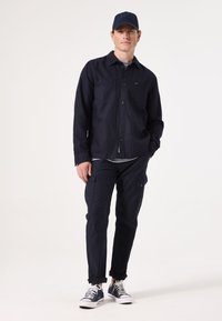 Marineblauwe button-up shirt met twee voorzakken, gecombineerd met bijpassende cargobroek. Versterkt met een marineblauwe pet en witte sneakers.