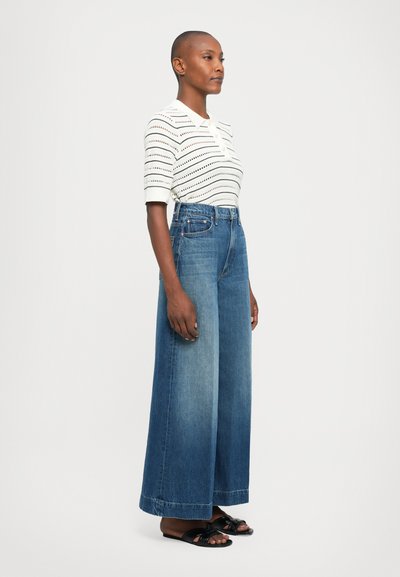 Femme se tenant debout dans un jean large bleu, un top en tricot blanc à manches courtes avec des rayures noires, et des sandales ouvertes noires sur un fond blanc.