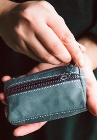 Kožená peňaženka v tlmenej zelenej farbe s čiernym zipsom a bielymi švy. Ruka otvára peňaženku.