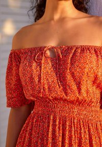 Femme portant une robe orange à épaules dénudées avec des manches courtes, un détail à nouer au niveau de la poitrine et une taille froncée.