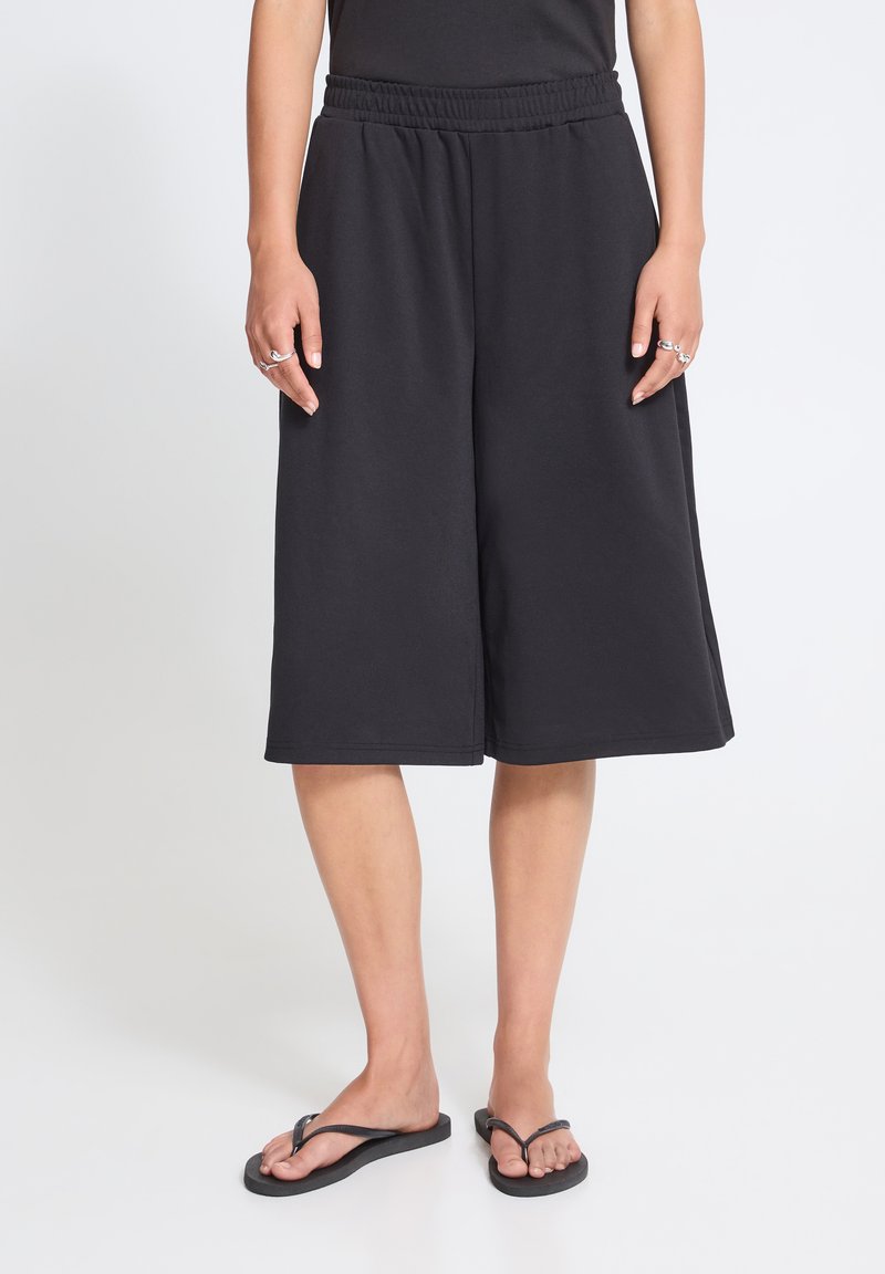 Personne portant un short noir large jusqu'aux genoux avec une taille élastique et des tongs noires, debout devant un fond uni.