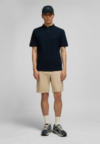 Maritimer Poloshirt in Marineblau mit Kragen, kombiniert mit beigen, hochgekrempelten Shorts. Abgerundet mit dunklen Sneakers und einer passenden Kappe, vor einem neutralen Hintergrund.