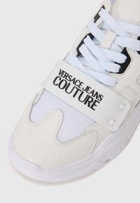Baskets blanches avec une tige en cuir texturé et des sections en mesh, dotées d'une sangle blanche portant le texte "VERSACE JEANS COUTURE" en noir.