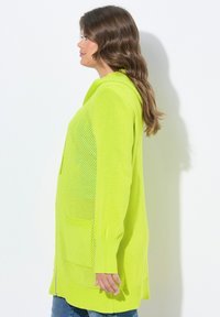 Cardigan lungo, di un vivace verde neon, con cappuccio e tasche laterali, caratterizzato da una texture a maglia e un design a rete aperta lungo i lati.