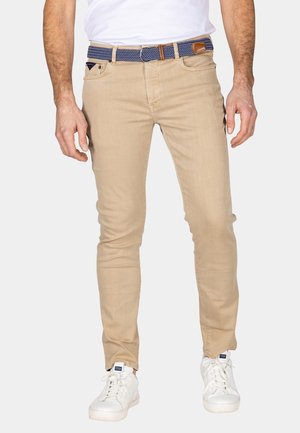 Beige slim-fit jeans met een geweven blauwe riem met bruine accenten, gedragen met witte sneakers, die de lichte denimtextuur benadrukken.