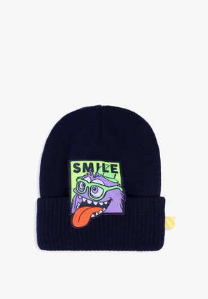 Bonnet tricoté bleu foncé avec un patch d'un monstre violet de dessin animé portant des lunettes vertes, langue tirée, et le mot "SMILE" au-dessus de sa tête.