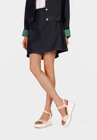 Sandali con zeppa in beige chiaro con tomaia testurizzata, punta aperta, cinturino alla caviglia e suola bianca a contrasto. Abbinati a una gonna in denim scura.