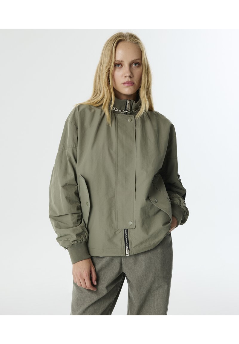 Giubbotto bomber verde oliva chiaro con colletto alto, zip nascosta e chiusure a pressione. Presenta polsini elastici e tasche laterali. Texture liscia.