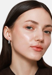 Orecchino a cerchio argentato in esposizione, caratterizzato da una superficie liscia e lucida. La modella ha capelli castani lisci, pelle chiara e un trucco delicato.