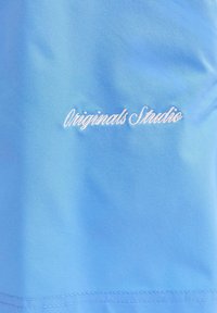 Tissu bleu clair avec texte brodé blanc "Originals Studio" centré près de la couture.