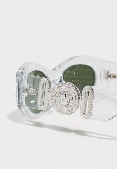 Ochelari de soare supradimensionați, transparenți, cu lentile verzi, având un accent decorativ din metal argintiu în formă de cap de leu pe latură.
