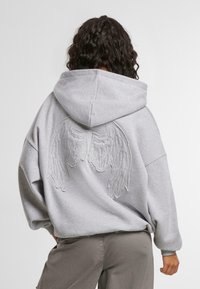 Graue, übergroße Kapuzenjacke mit einem großen, gestickten Flügeldesign auf dem Rücken, ausgestattet mit einer Kordelzugkapuze und einer lässigen Passform. Weicher Stoff.