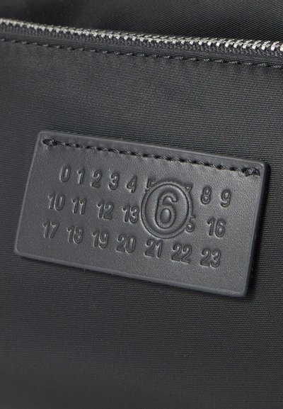 Juodas odinis pleistras su įspaustomis skaičiais nuo 0 iki 23, apvaliu skaičiumi 6, siuvamas ant juodos medžiagos šalia sidabrinio užtrauktuko.