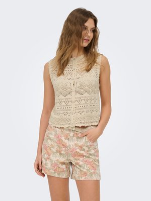 Kvinde iført beige hæklet ærmeløs top og blomsterprintede shorts, stående med den ene hånd i lommen og kigger let til siden.