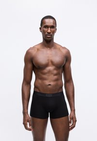 Hombre musculoso de pie, mirando de frente, en calzoncillos negros de la marca Hugo, contra un fondo blanco liso.