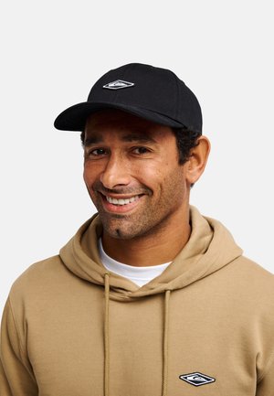 Man die lacht, draagt een beige hoodie en een zwarte pet, beide met bijpassende wit-zwarte logo's in diamantvorm.