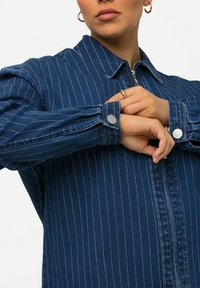 Blå denimjacka med vita vertikala ränder, spetskrage och tryckknappsmuddar. Tyget har en slät yta och en figurnära design.