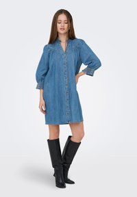 Νεαρή γυναίκα που φοράει μπλε τζιν φόρεμα με κουμπιά και φουσκωτά μανίκια, καθώς και μαύρες μπότες μέχρι το γόνατο, στέκεται με το ένα χέρι στη μέση.