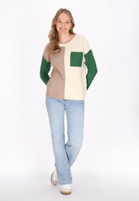 Strickpullover mit cremefarbenen, grünen und beigen Abschnitten; verfügt über eine strukturierte grüne Tasche; getragen mit hellblauen Jeans und weißen Sneakers.