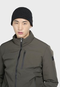 Giacca verde oliva con zip, colletto alto e dettagli neri. Beanie nero a coste indossata dal modello, vista ravvicinata del volto.