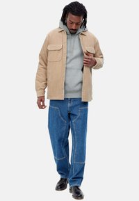 Chaqueta de camisa de pana marrón clara con dos bolsillos en el pecho, superpuesta sobre una sudadera gris claro; jeans anchos azules con detalles de costura visibles; zapatos negros.