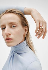 Se llevan pendientes en forma de lágrima de oro y un anillo de multipes de piedras. El modelo tiene el cabello largo, un cuello de tortuga azul claro y un fondo neutro.