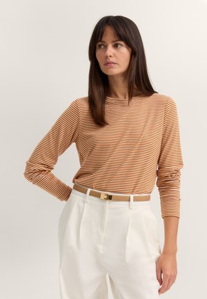Woman with long dark hair wearing a brown and white striped long-sleeve shirt tucked into white pants with a beige belt, looking sideways.