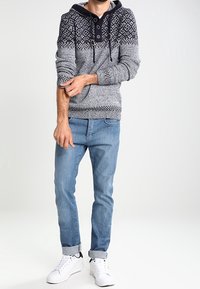 Gestrickter grauer und schwarzer Pullover mit Muster und Kapuze, mit einem knöpfbaren Ausschnitt, kombiniert mit hellblauen, hochgekrempelten Jeans und weißen Turnschuhen.