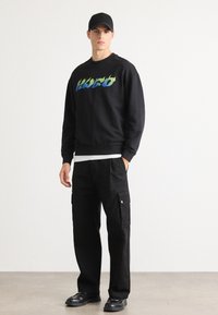 Young man wearing black cap, black sweatshirt with blue-green "HUGO" text, black cargo pants, and black shoes standing on gray floor.
