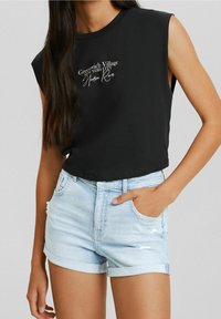 Schwarzes ärmelloses Crop-Top mit weißem Text, kombiniert mit hellblauen Jeansshorts mit fransigen Kanten und umgeschlagenen Säumen.