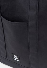Zwarte stoffen tas met een voorvak, stevige stiksels en een witte Timberland logo patch. Eenvoudig ontwerp met een textuur oppervlak.