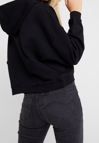 Sudadera negra corta con capucha y puños acanalados, combinada con unos jeans oscuros ajustados de color gris que presentan detalles de costura y una etiqueta de logo.