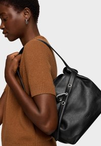 Czarna skórzana torba na ramię o fakturowanym wykończeniu, z zapięciem na ściągacz i srebrnymi akcentami, noszona na brązowym swetrze.