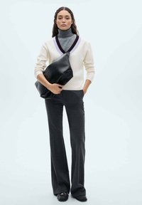 Creme V-ringad stickad tröja med grå och lila detaljer, grå polotröja, mörkgrå vida byxor, håller en svart mjuk läderclutch.