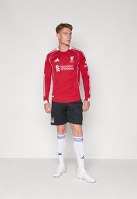 Rød langærmet fodboldtrøje med hvide accenter og logo, parret med sorte shorts, hvide strømper med blå striber og hvide fodboldstøvler.
