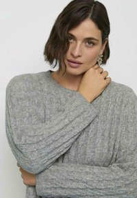 Maglione grigio a maglia con texture a coste e maniche ampie, presentato con una mano che poggia sul colletto, evidenziando il design accogliente.